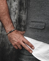 Close-up of a person wearing a dark gray textured waistcoat over a white shirt, with one hand resting on the waistline and a beaded bracelet visible on the wrist.