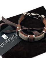 Brown leather and metal bracelet on a dark surface with 'otherwise jewelry' card.