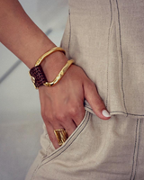 A close-up of a person's hand wearing three gold-toned jewelry pieces: a thick textured bangle, a thinner smooth bangle, and a wide rectangular band ring, paired with a beige linen top.