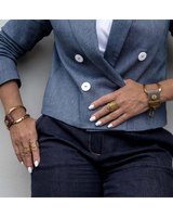 A close-up of a person wearing a slate blue double-breasted blazer with white buttons, paired with dark jeans and accessorized with gold rings and bracelets.