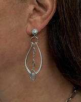 A close-up of a person's ear wearing a silver teardrop dangle earring with a small dangling charm, against skin and dark brown hair.
