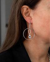 Elegant loop earrings feature a large silver circle with a smaller oval and an Austrian crystal pendant, worn by a woman.