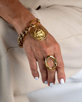 A close-up of a person's hand wearing a chunky gold chain bracelet with a large circular gold coin medallion and a wide gold ring, set against a soft beige linen background.