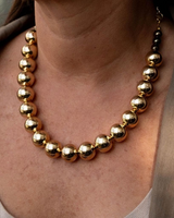 A close-up of a gold beaded statement necklace worn by a model, featuring large polished metallic gold spheres.