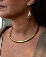 A close-up of a woman wearing a textured gold snake chain necklace and a matching gold teardrop dangle earring.