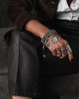 Chunky coin charm bracelet with leather closure, featuring a heavy curb chain and antique coins, worn on a wrist with silver rings.