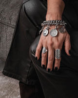 Chunky silver coin charm bracelet with leather closure, worn with layered rings, showcasing bold, vintage-inspired pieces.