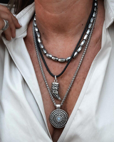 Layered leather and metal necklaces set worn with a loose, collared shirt. Features black leather cord, metallic accents, and blue stone.