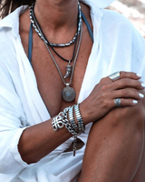 Layered leather and metal necklaces featuring black leather cords, silver accents, and a blue stone pendant, worn over a white top.