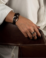 Unisex leather wrap bracelet in dark brown with a silver o-ring cuff, worn on a wrist with minimalist rings.