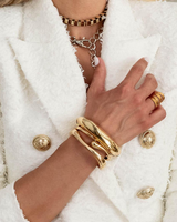 Chunky gold cuff bangles stacked on a model's wrist, styled with a white blazer and layered necklaces.