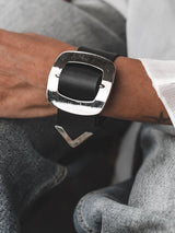 Wide black leather bracelet featuring a large silver-plated buckle, worn on the wrist with a chunky strap.