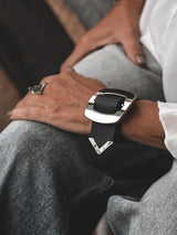Wide black leather bracelet featuring a large silver buckle, worn as a chunky cuff on a wrist.