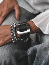 Wide black leather bracelet with a large silver buckle, worn with a stacked silver bead bracelet on a hand resting on denim.