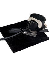 Wide black leather bracelet featuring a large silver buckle, presented on a soft black pouch with a ribbon closure.