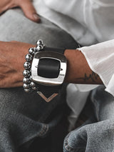 Wide black leather cuff bracelet featuring a large silver buckle, worn with a silver bead bracelet on an arm.