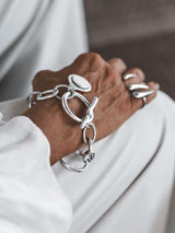 Silver Phaistos Disc Bracelet featuring a teardrop pendant and asymmetric chain, worn on a wrist with a light, casual outfit.