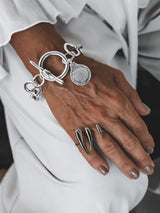 Silver Phaistos Disc bracelet features an asymmetric link chain, large toggle closure, and teardrop pendant, worn on a wrist.