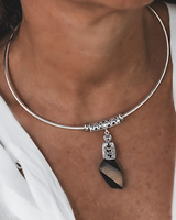 A close-up of a person wearing a thin silver bangle necklace with a dangling pendant. The pendant features a silver filigree cap and a faceted dark stone charm with a beige center.
