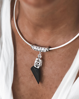 A close-up of a person wearing a silver-toned bangle necklace with an ornate cylindrical bead, a filigree charm, and a dangling black triangular pendant.