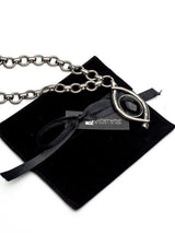 A silver-toned chain necklace with a stylized eye pendant containing a black center, displayed on a black velvet pouch with a black ribbon and a visible 'Mutherwise' brand tag.
