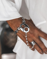 A close-up of a person's hand wearing a chunky silver chain bracelet with a large circular disc and smaller chain links, styled with a silver ring, against a white garment background.