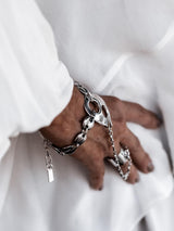 A close-up of a person's hand and wrist wearing a chunky silver chain bracelet with large interlocking links and a small dangling tag, styled with a white sleeve and white fabric background.