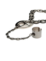 A silver-toned metal chain bracelet featuring a large oval medallion, a short chain segment, and a cylindrical ring charm, shown on a plain white background.