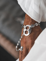 A close-up of a person's hand wearing a chunky silver chain bracelet with a large oval cutout charm and a matching silver ring on the finger, styled with a white sleeve.