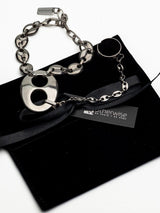 A silver-toned metal chain bracelet with a large circular O-shaped charm and a small oval charm, displayed on a black velvet pouch with a black ribbon and a branded tag reading 'otherwise by Sarah x Elvyx'.