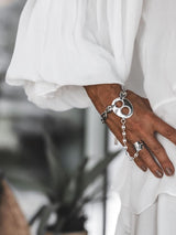A close-up of a person's hand wearing a silver chain bracelet with a large circular padlock charm and a silver ring, styled with a flowing white dress.