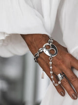 A close-up of a person's hand wearing a chunky silver chain bracelet with a large circular cutout pendant and a matching chain draped over the fingers, paired with a silver ring, against a white draped garment.