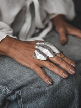 Chunky, sculptural knuckle ring in irregular, textured zinc alloy; worn on a hand resting on denim fabric.