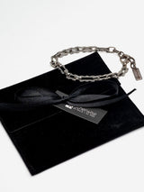 Stainless steel rectangular link chain anklet resting on a black velvet pouch with a ribbon, featuring a chunky, minimalist design.