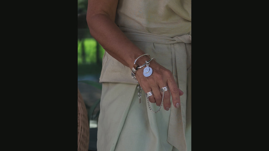 A close-up of a person's hand wearing multiple silver-toned jewelry pieces, including a wide cuff bracelet with a disc charm, a chain bracelet, and several rings, paired with a light beige garment.