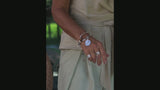 A close-up of a person's hand wearing multiple silver-toned jewelry pieces, including a wide cuff bracelet with a disc charm, a chain bracelet, and several rings, paired with a light beige garment.
